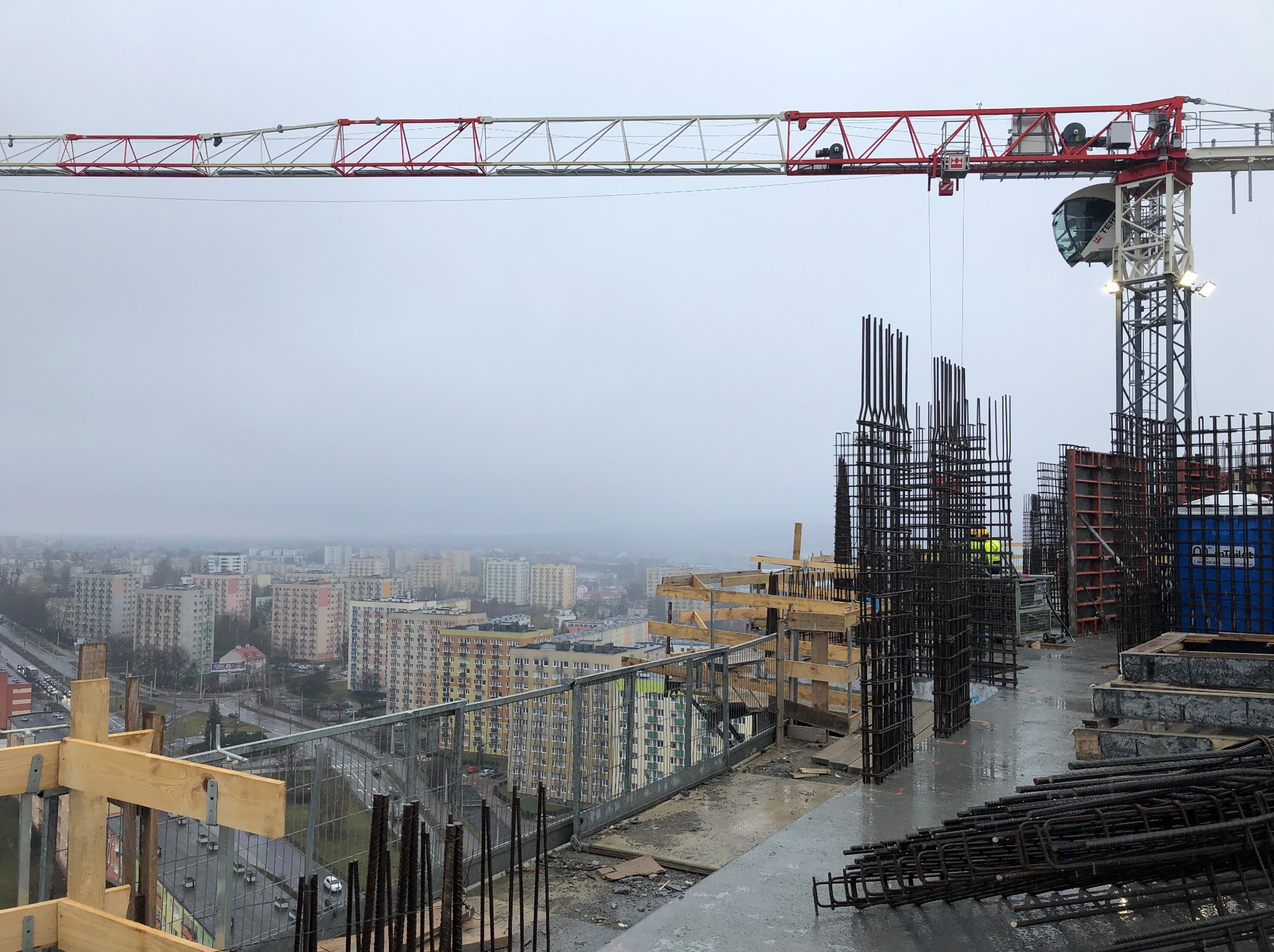 Budowa budynku żelbetowego w deszczowy dzień: widok z góry na zbrojenie, szalunki i dźwig na tle miejskiej zabudowy w oddali, pochmurne niebo.