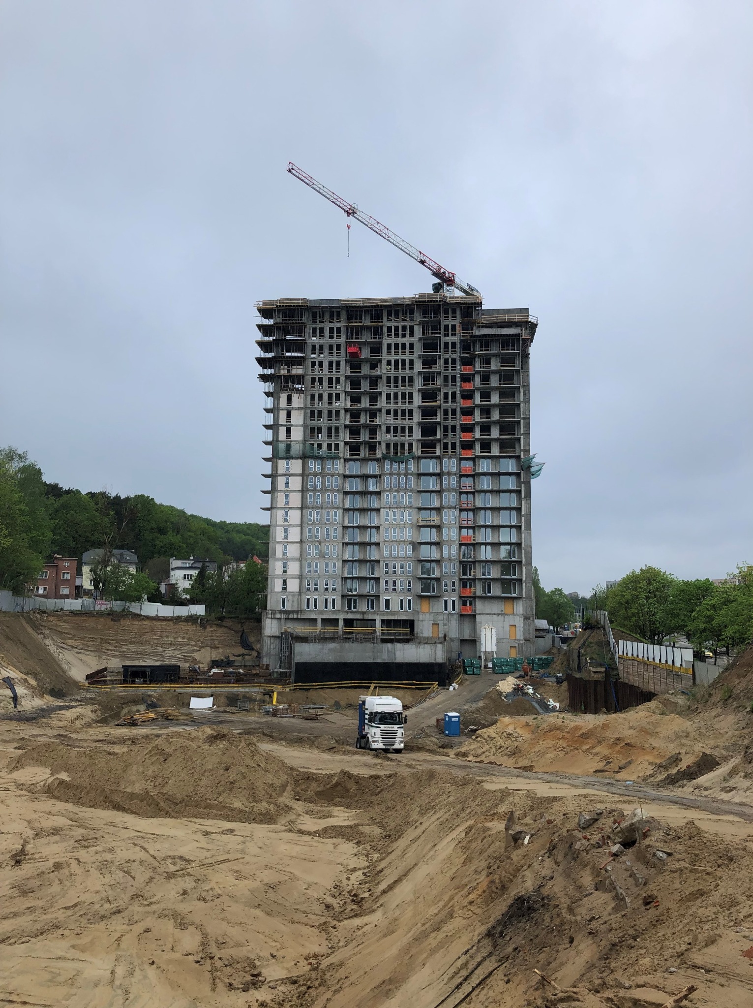 Wysoki budynek żelbetowy w trakcie budowy, widoczny żuraw budowlany, ciężarówka i wykop pod fundamenty. Tło stanowią drzewa i pochmurne niebo.