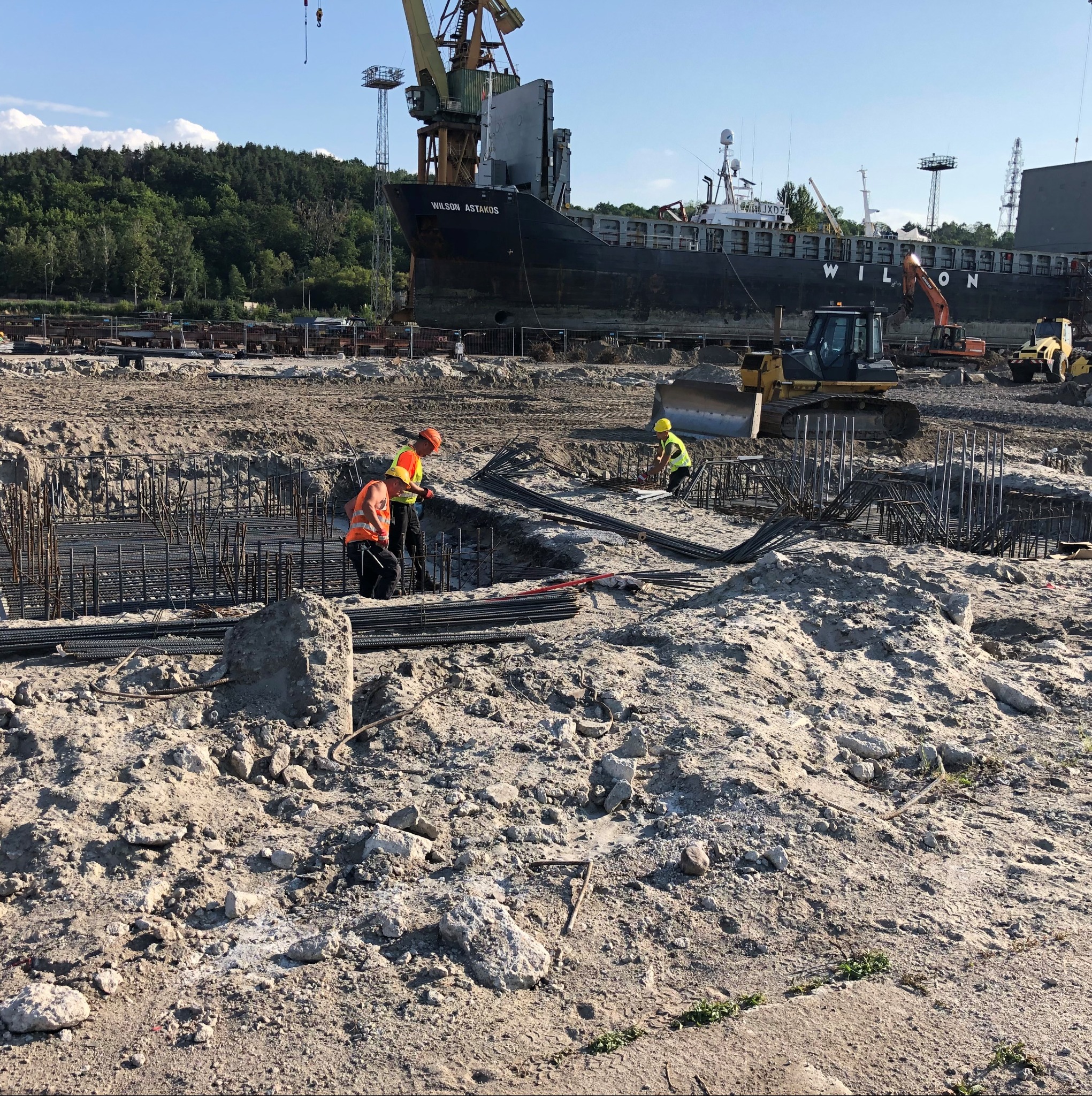 Praca zbrojarska na placu budowy w porcie: zbrojenie fundamentów, pracownicy w kamizelkach, ciężki sprzęt i statek Wilson Astakos w tle, słoneczny dzień.