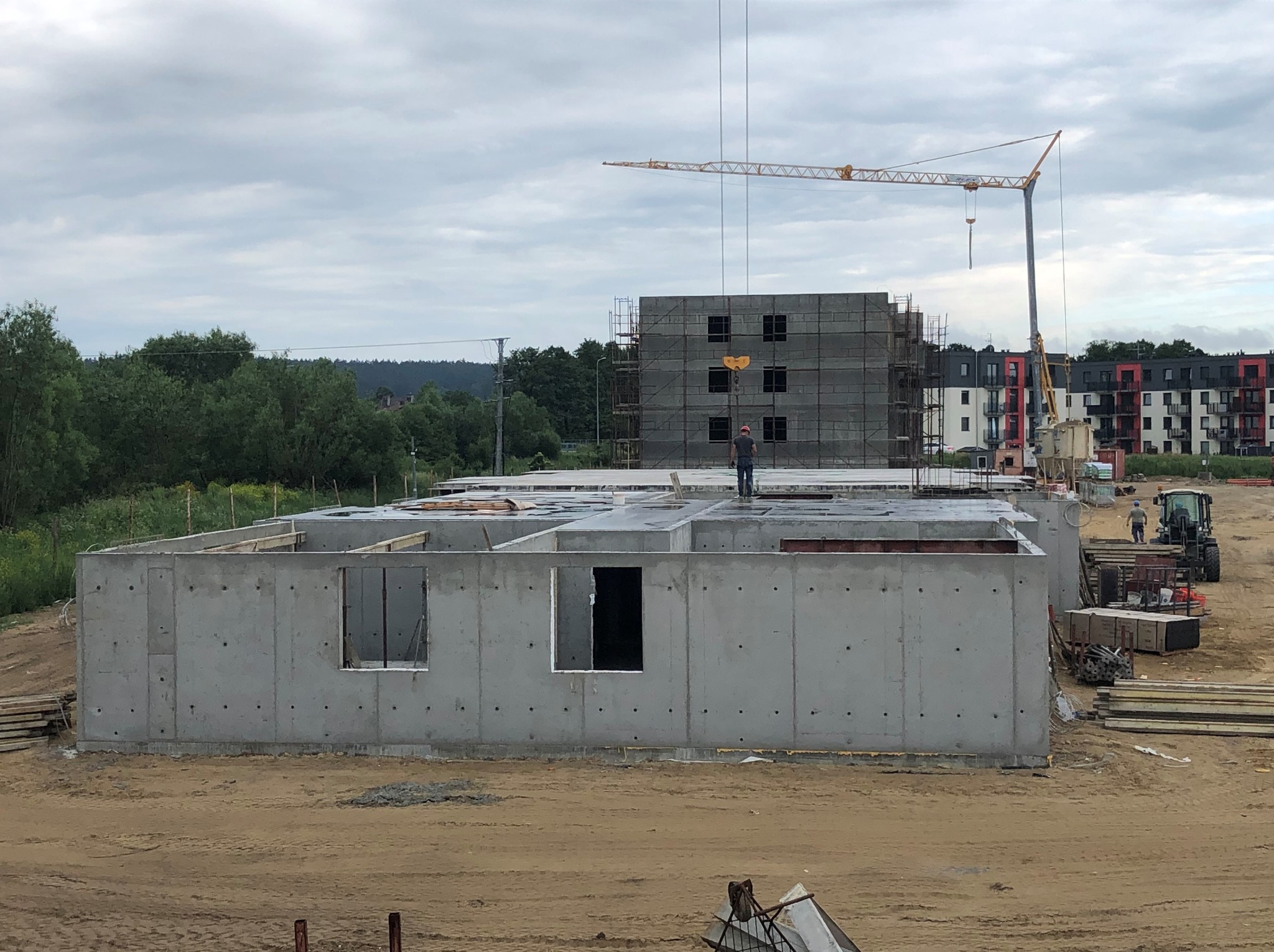 Surowy stan budynku żelbetowego z widocznym fundamentem i ścianami, na tle placu budowy z dźwigiem i budynkiem w rusztowaniach. Robotnik na dachu.