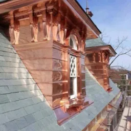 Miedziane obróbki blacharskie lukarn na dachu pokrytym łupkiem, widoczne detale architektoniczne i rynna. Dach w klasycznym stylu, na tle błękitnego nieba.