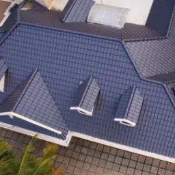Widok z góry na ciemnoszary dach z dachówki ceramicznej, z licznymi lukarnami i białymi obróbkami blacharskimi. W tle widoczny fragment ogrodu i chodnika z kostki.