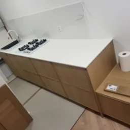 Nowoczesna, jasna kuchnia w trakcie montażu: drewniane szafki, biały blat z wbudowaną płytą grzewczą i zlewem. Widoczne elementy instalacji elektrycznej.