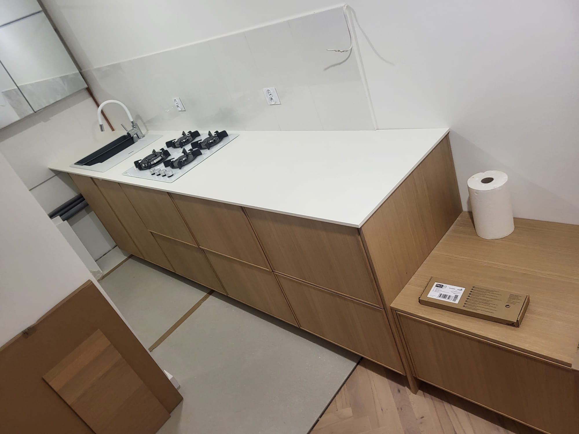 Nowoczesna, jasna kuchnia w trakcie montażu: drewniane szafki, biały blat z wbudowaną płytą grzewczą i zlewem. Widoczne elementy instalacji elektrycznej.