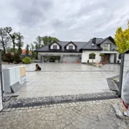 Ułożona kostka granitowa na podjeździe do domu, widoczny pies siedzący na kostce, fragment niezakończonej pracy przy bramie wjazdowej i osoba w kadrze.