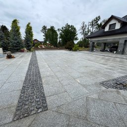 Prof-bruk - Wykończony podjazd z szarej kostki granitowej, z ciemniejszym pasem z kostki brukowej, prowadzący do domu z garażem. W tle zieleń i drzewa.