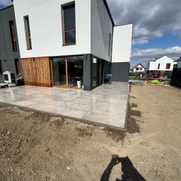 Prof-bruk - Nowoczesny taras z szarych płyt na gruncie przy bliźniaku z drewnianą fasadą. Widoczne sąsiednie domy i fragment nieba z chmurami.