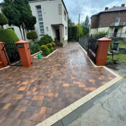 Ukończony podjazd z kostki brukowej w odcieniach brązu i pomarańczy, obramowany jasną kostką. Widoczne zadbane rośliny i fragmenty budynków w tle.