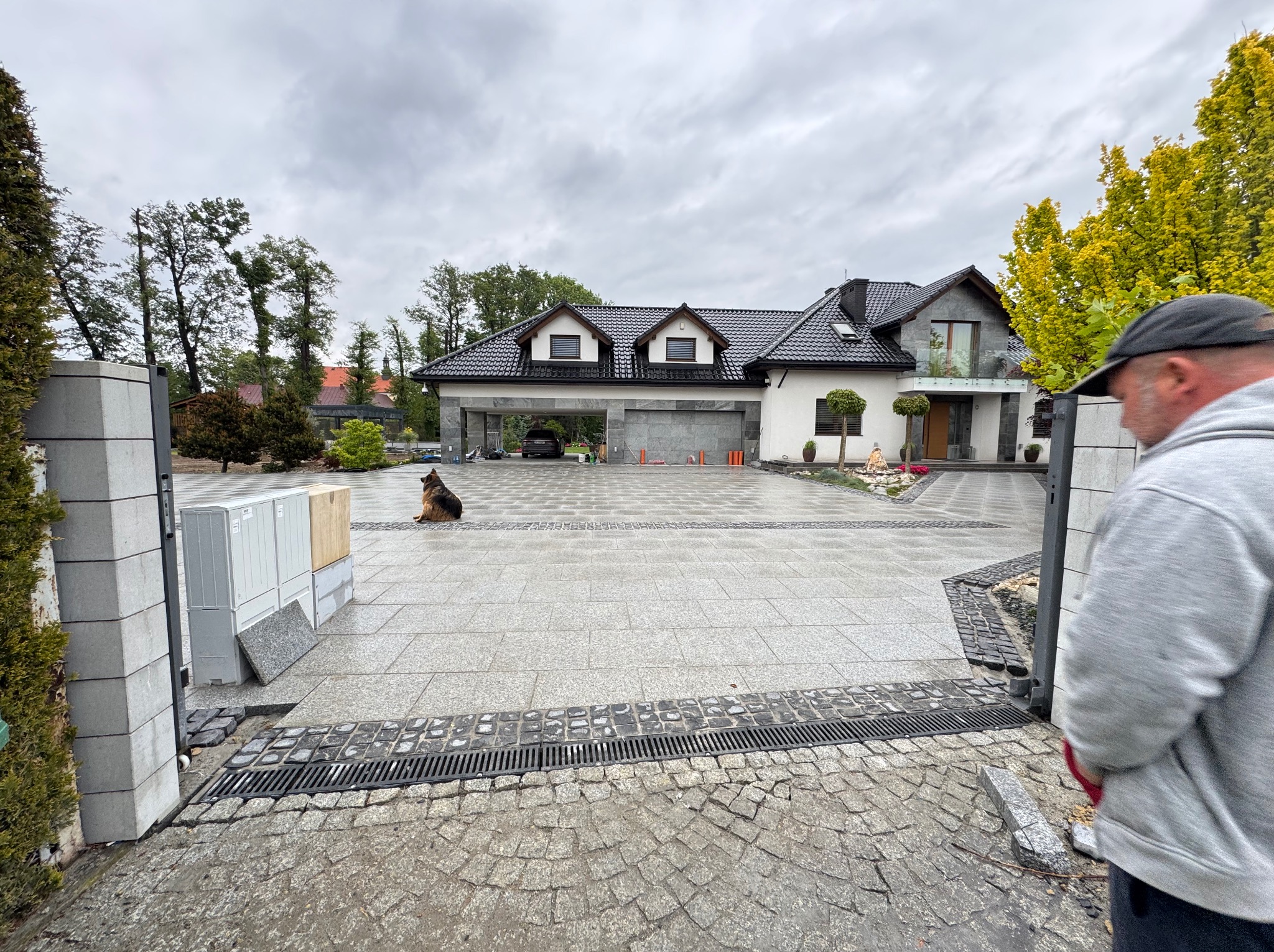 Ułożona kostka granitowa na podjeździe do domu, widoczny pies siedzący na kostce, fragment niezakończonej pracy przy bramie wjazdowej i osoba w kadrze.