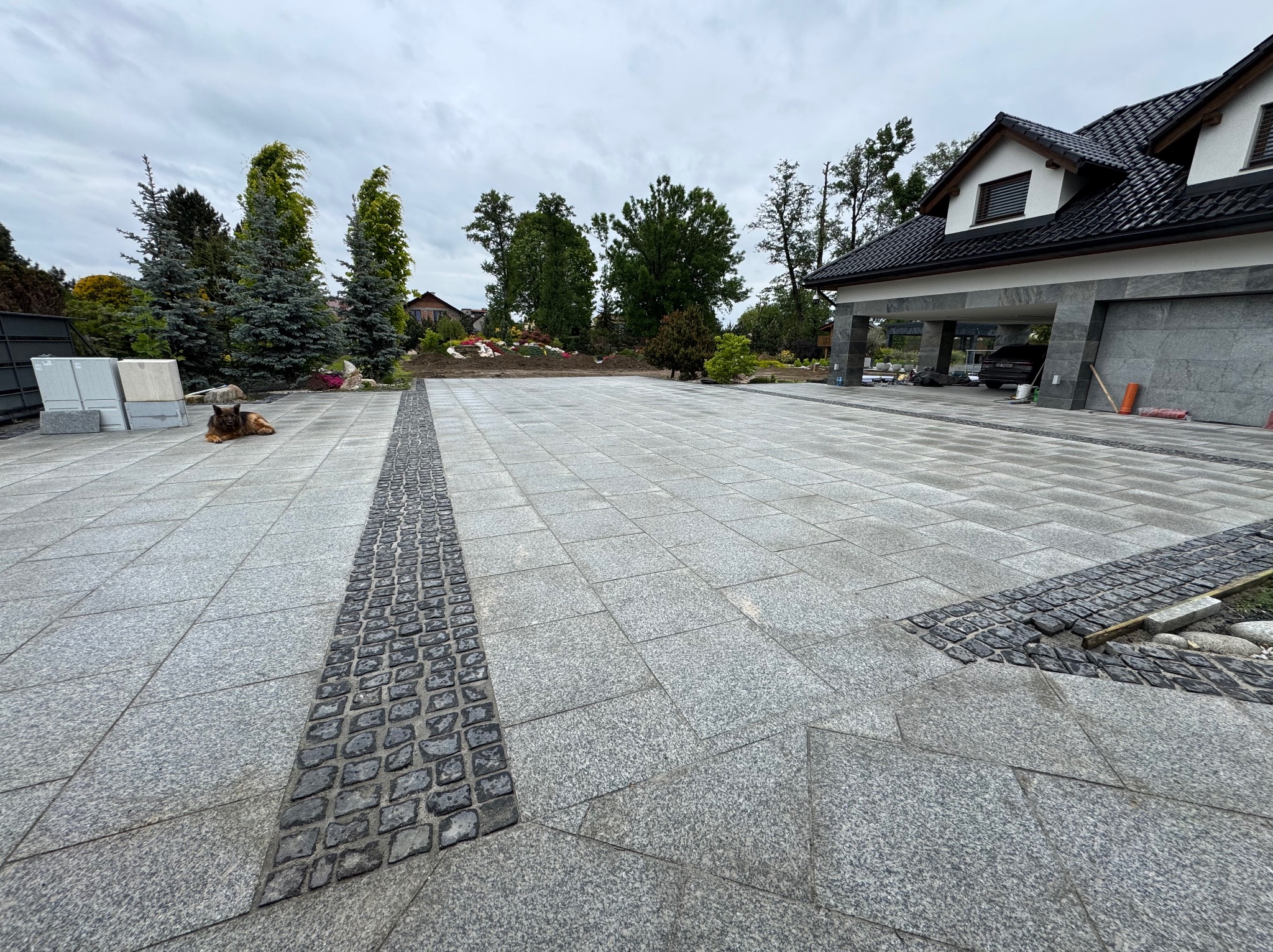 Wykończony podjazd z szarej kostki granitowej, z ciemniejszym pasem z kostki brukowej, prowadzący do domu z garażem. W tle zieleń i drzewa.