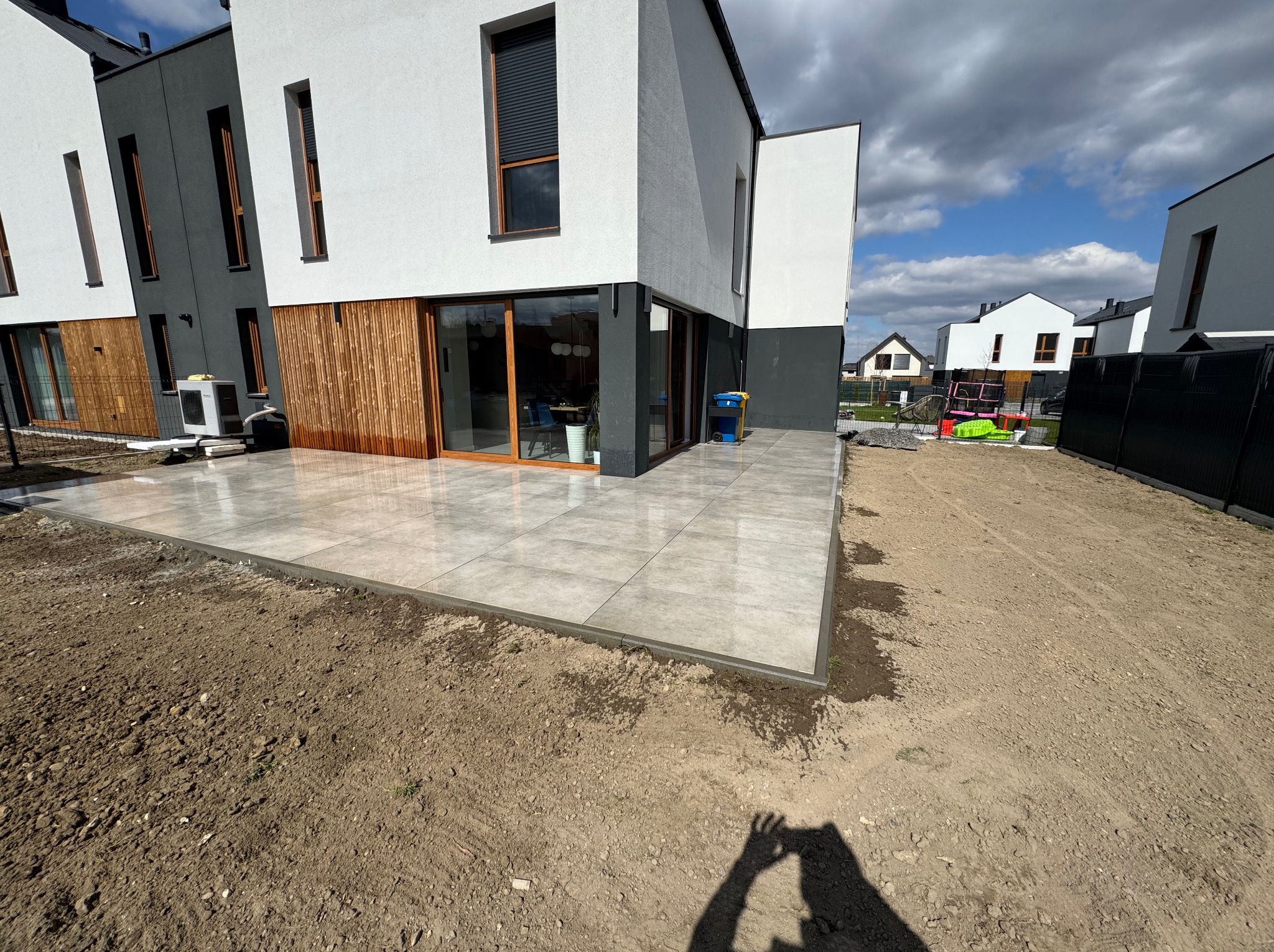 Nowoczesny taras z szarych płyt na gruncie przy bliźniaku z drewnianą fasadą. Widoczne sąsiednie domy i fragment nieba z chmurami.