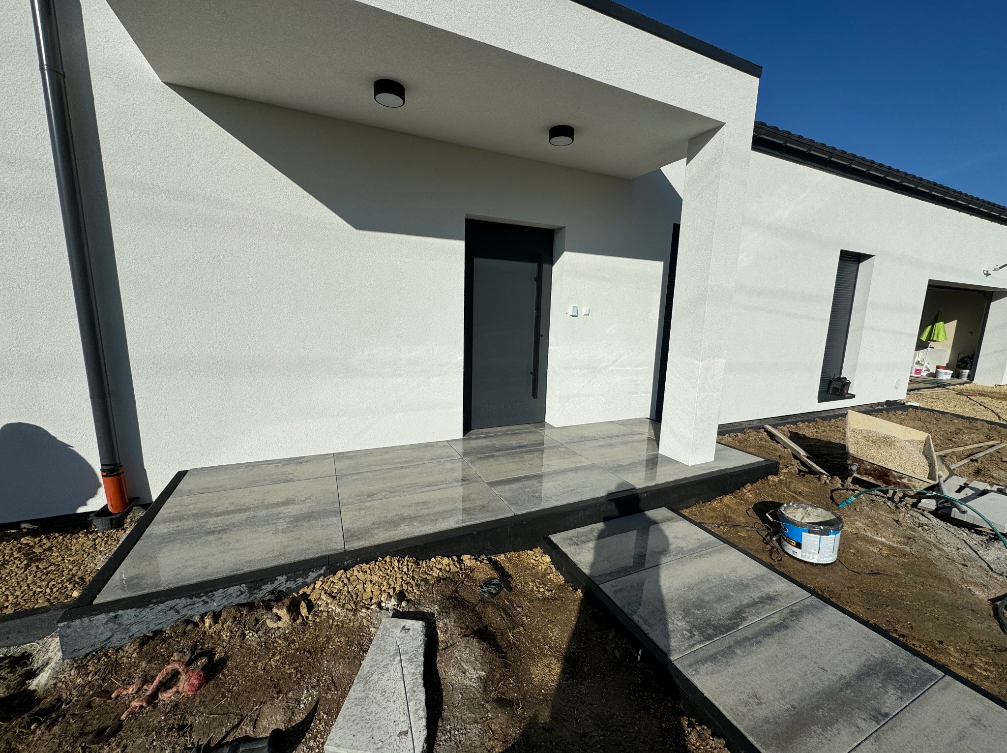 Nowoczesne wejście do domu w Jawiszowicach z szarych płyt. Ułożona kostka brukowa prowadzi do drzwi. Widoczne prace wykończeniowe wokół.