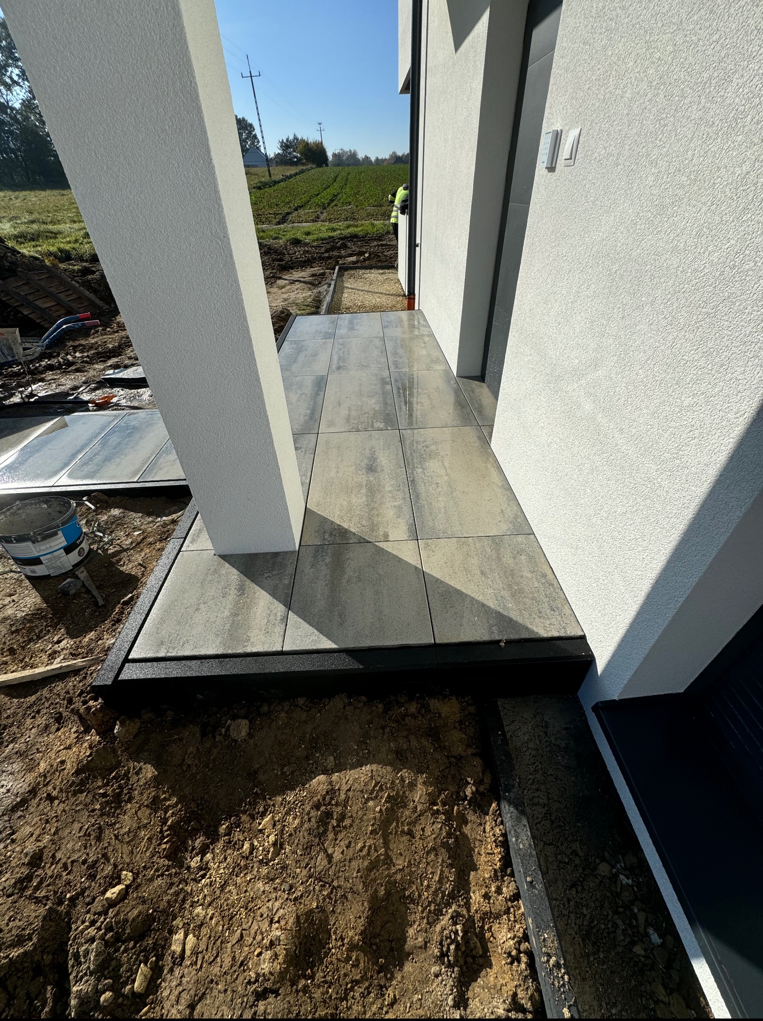 Nowoczesny taras z szarych płyt betonowych przy budynku z białą elewacją. Widoczny fragment ogrodu i słup podtrzymujący zadaszenie. Prace w toku, widoczne narzędzia i ziemia.