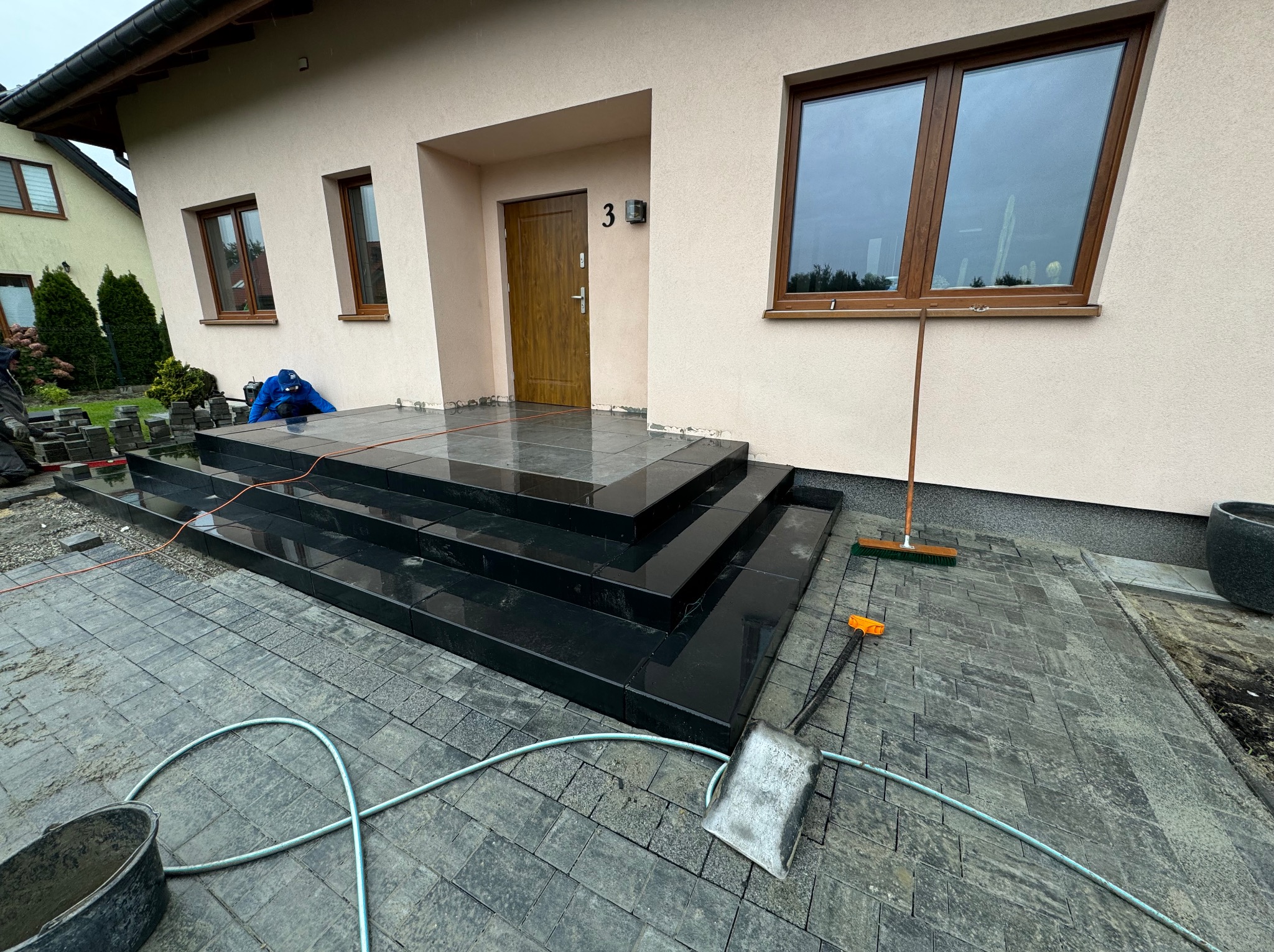Nowoczesne, czarne schody z kostki, prowadzące do domu z numerem 3. Widoczna ułożona kostka brukowa, narzędzia pracy: łopata, szczotka, wiadro i wąż ogrodowy.