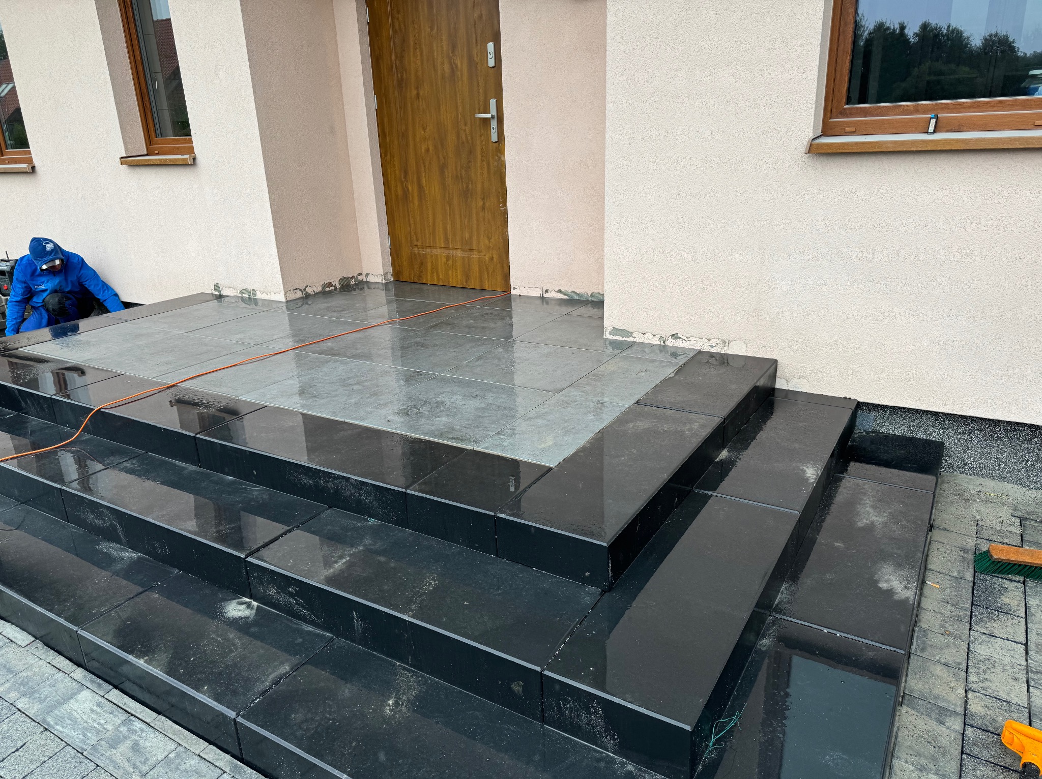 Schody wejściowe wyłożone błyszczącymi, czarnymi płytkami. W tle pracownik w niebieskim kombinezonie. Widoczne detale: fuga, faktura płytek, elewacja budynku.