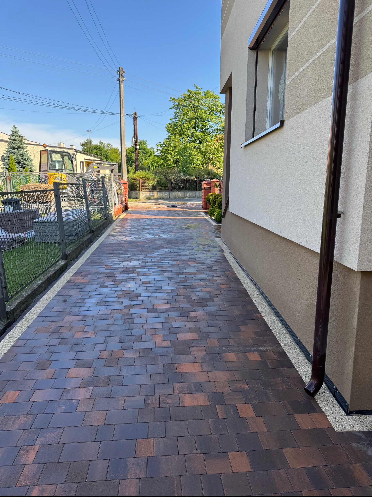 Ułożona, brązowo-grafitowa kostka brukowa tworząca ścieżkę wzdłuż domu, z widocznym fragmentem ogrodu i sprzętu budowlanego w tle. Kąt ujęcia podkreśla perspektywę.
