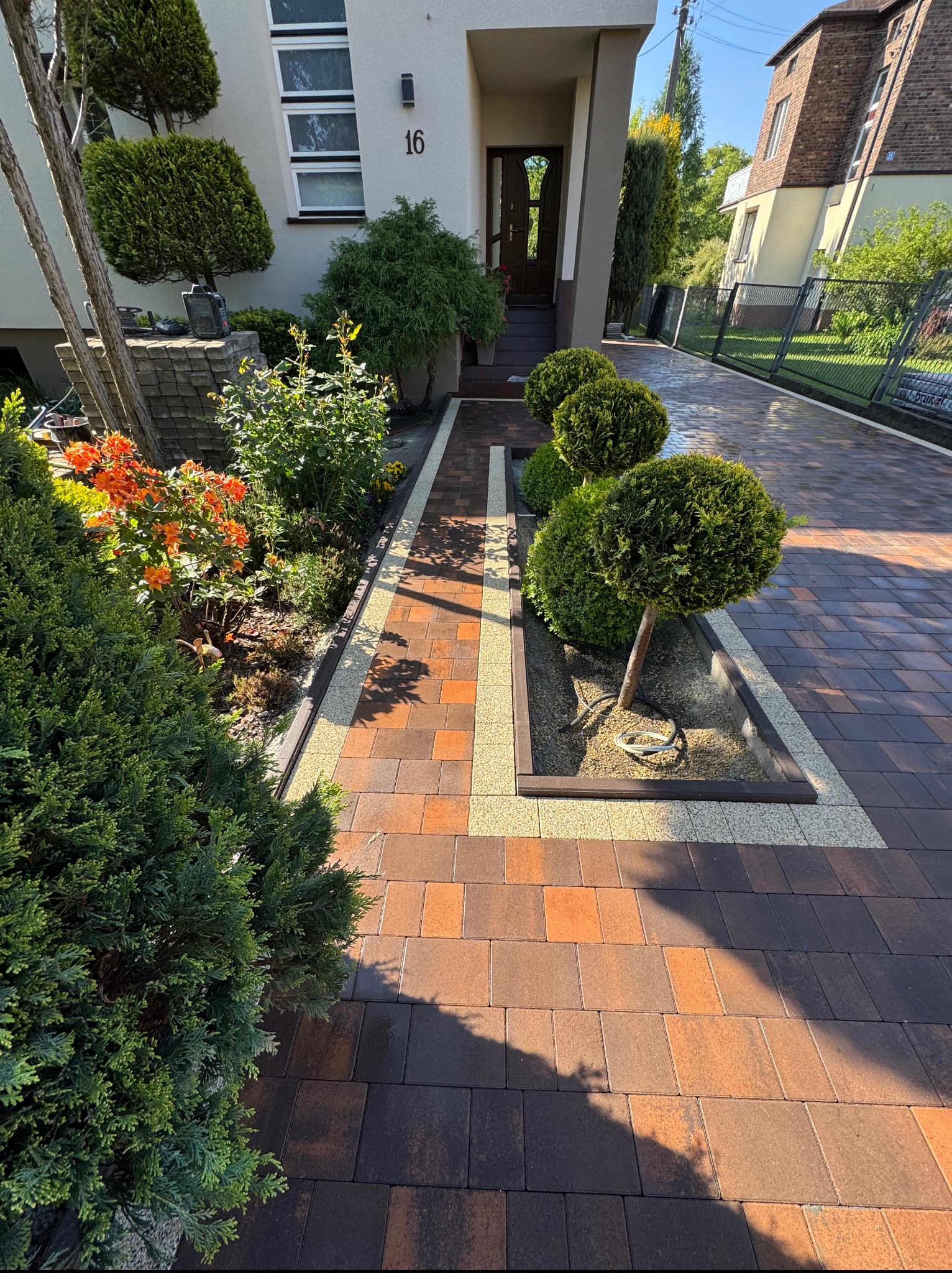 Ogród z alejką z kostki brukowej w odcieniach brązu i beżu, z równo przyciętymi krzewami i rabatami kwiatowymi. Widok na dom z numerem 16 w tle.