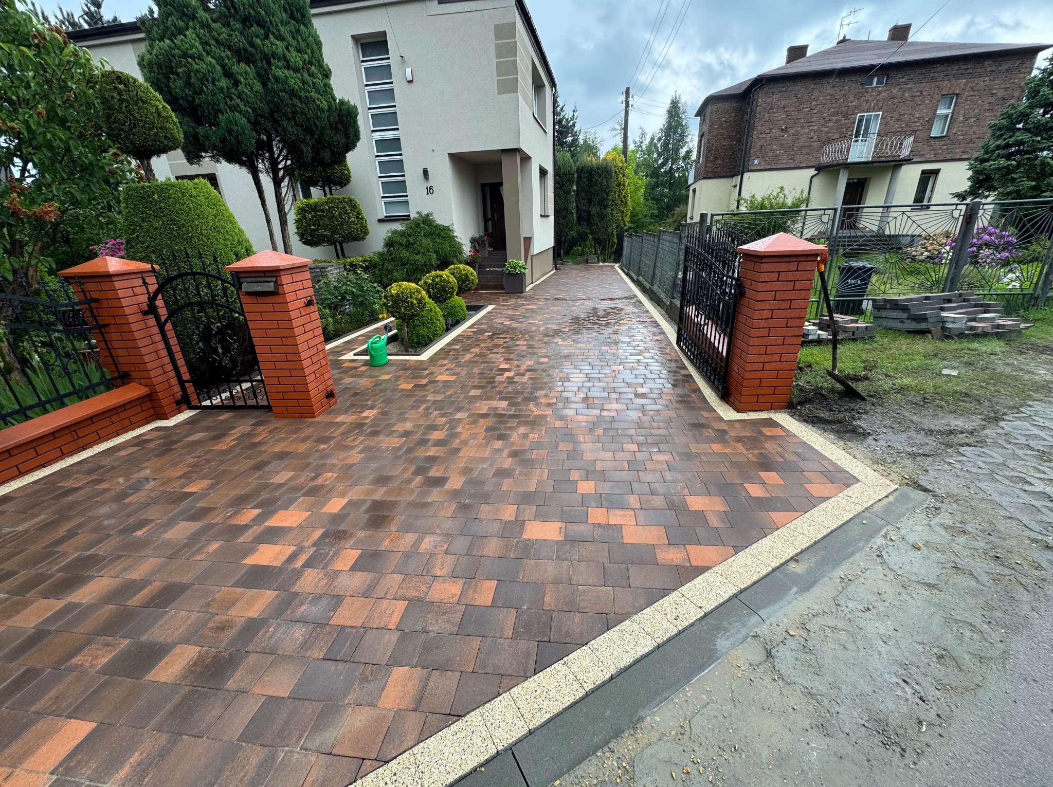 Ukończony podjazd z kostki brukowej w odcieniach brązu i pomarańczy, obramowany jasną kostką. Widoczne zadbane rośliny i fragmenty budynków w tle.