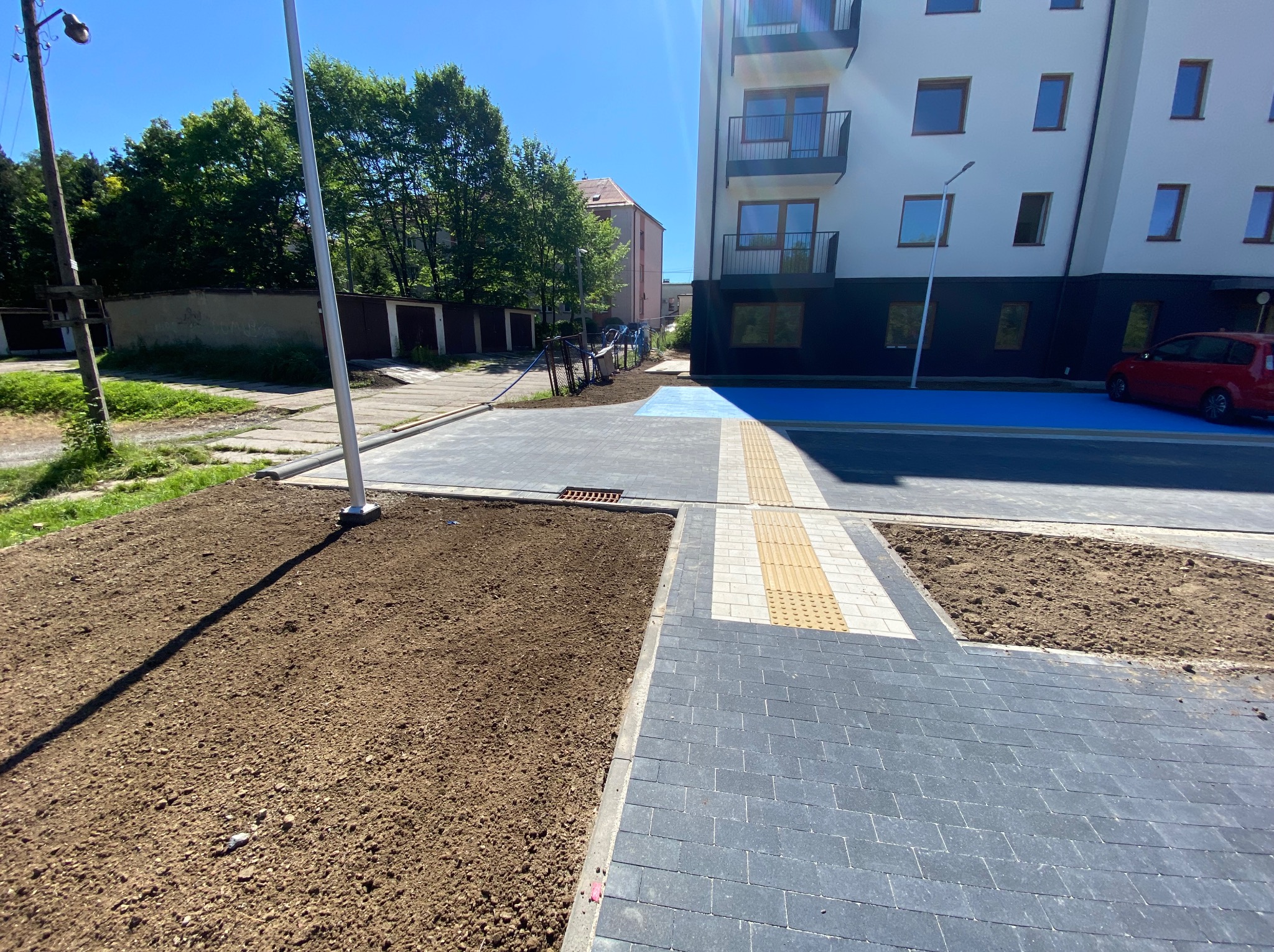 Nowa nawierzchnia z kostki brukowej przed budynkiem mieszkalnym, z elementami dla osób niewidomych. Przygotowane rabaty ziemne czekają na nasadzenia.