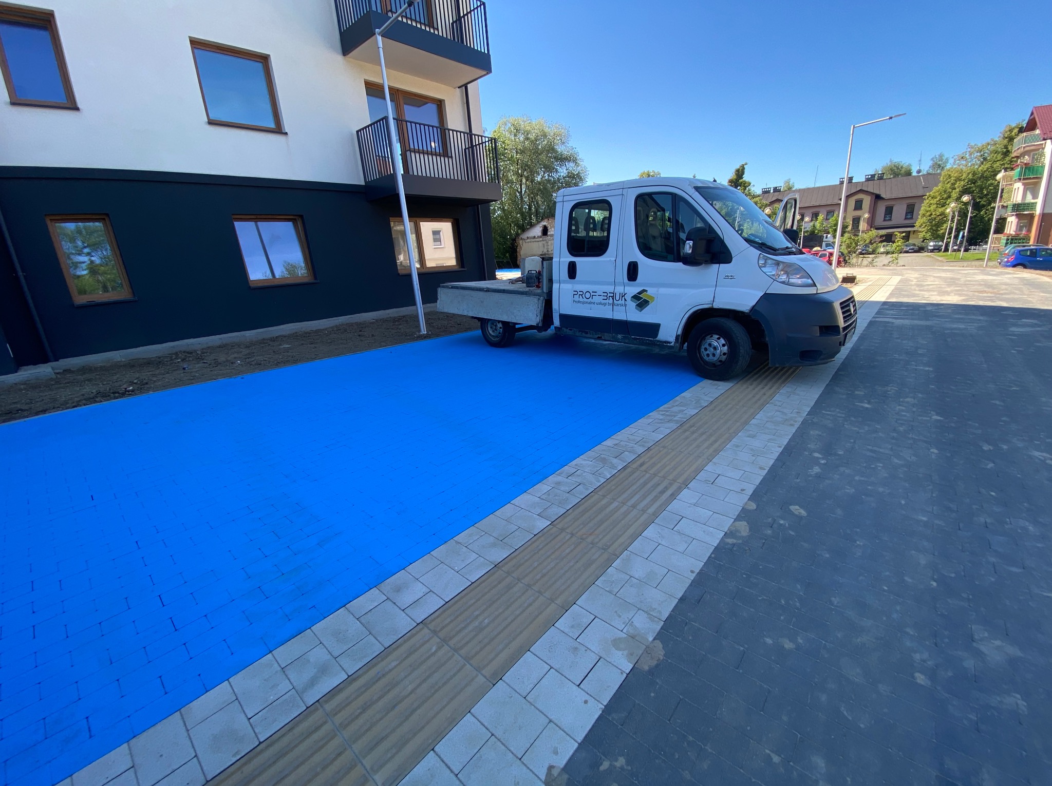 Niebieska kostka brukowa przy budynku z szarym pasem dla niewidomych i szarym brukiem. Biały bus firmy PROF-BRUK. Słoneczny dzień.