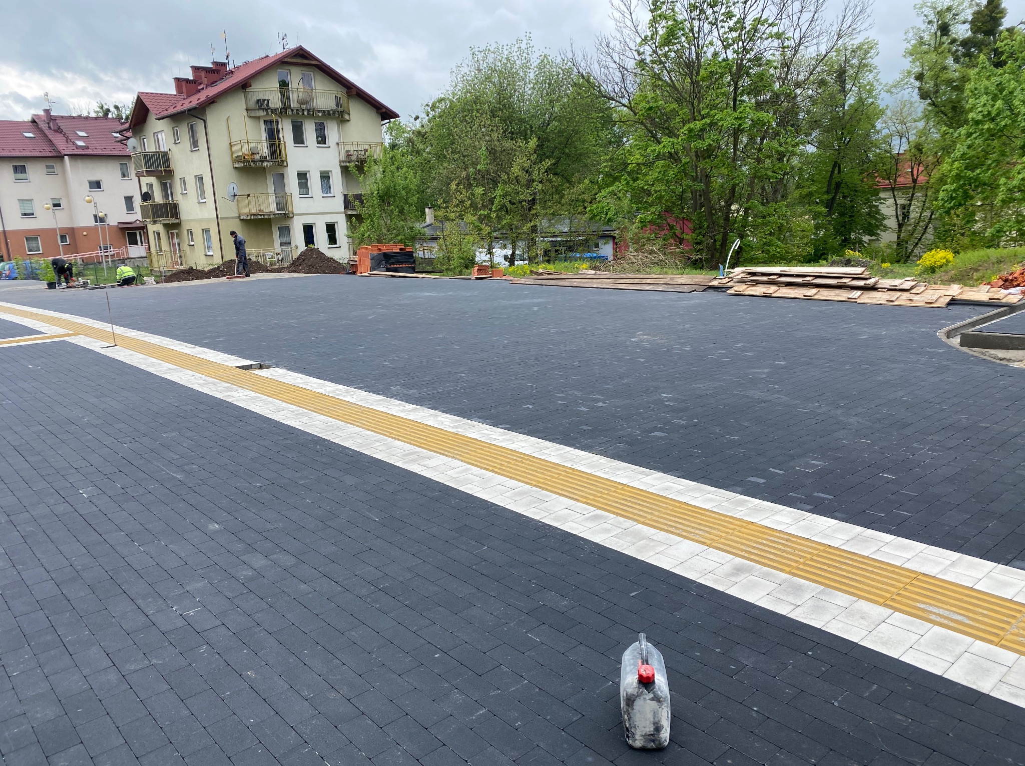 Ułożona, grafitowa kostka brukowa z żółtymi pasami dla niewidomych; w tle budynki i zieleń. Na pierwszym planie kanister. Widoczni pracownicy w oddali.