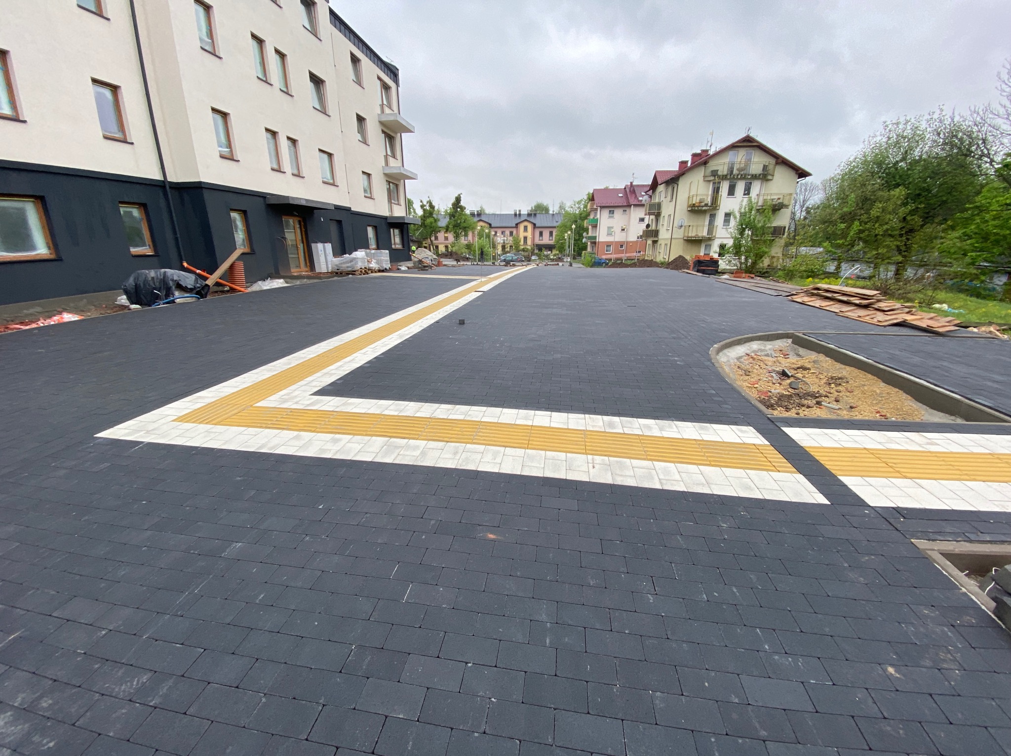 Ciemna kostka brukowa z żółtymi i białymi pasami dla niewidomych, przed budynkiem. Częściowo ułożona, widoczne materiały budowlane. Pochmurne niebo.