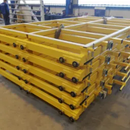 Stos żółtych, spawanych konstrukcji metalowych z rolkami, ułożonych w hali produkcyjnej. Elementy przygotowane do dalszego montażu. Widoczni pracownicy w tle.