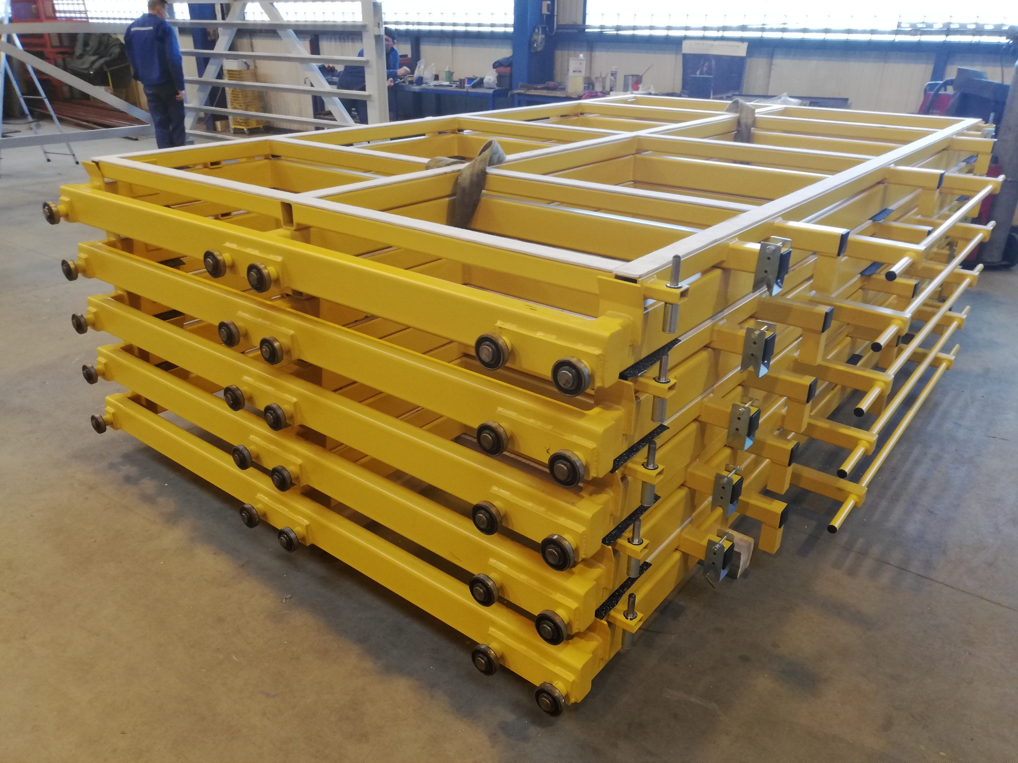 Stos żółtych, spawanych konstrukcji metalowych z rolkami, ułożonych w hali produkcyjnej. Elementy przygotowane do dalszego montażu. Widoczni pracownicy w tle.