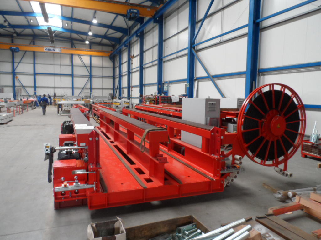 Czerwona, stalowa konstrukcja w hali produkcyjnej z suwnicą. Widoczne elementy złączne i niebieskie elementy konstrukcyjne hali.