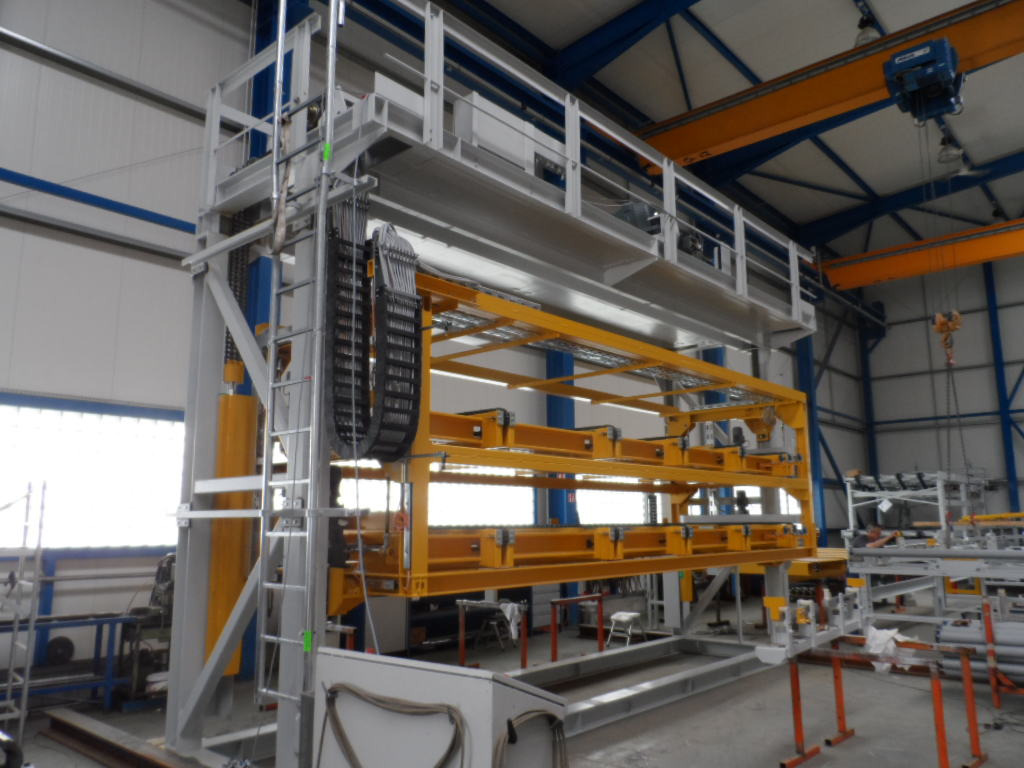 Wnętrze hali produkcyjnej z konstrukcją stalową w kolorze żółtym i srebrnym, widoczny suwnica pod stropem. Elementy konstrukcyjne w trakcie montażu.