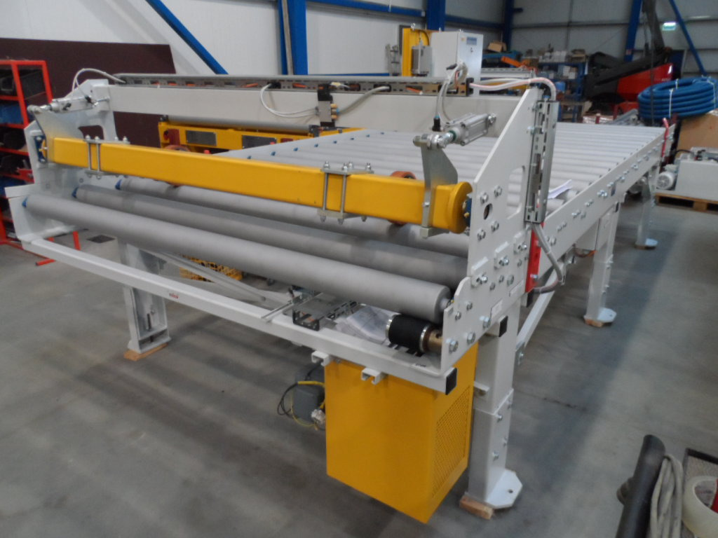 Detaliczny widok na białą, metalową konstrukcję z żółtymi akcentami, prawdopodobnie element linii produkcyjnej w hali. Widoczne rolki i mechanizmy.