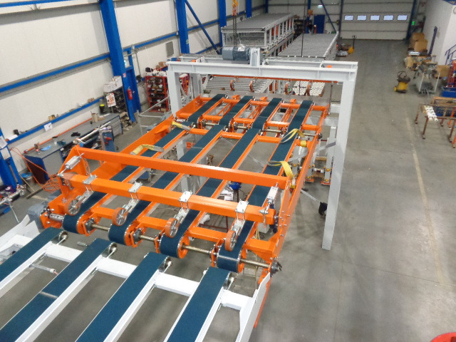Widok z góry na pomarańczowo-białą konstrukcję z pasami transportowymi w hali produkcyjnej; w tle inne maszyny i wyposażenie.
