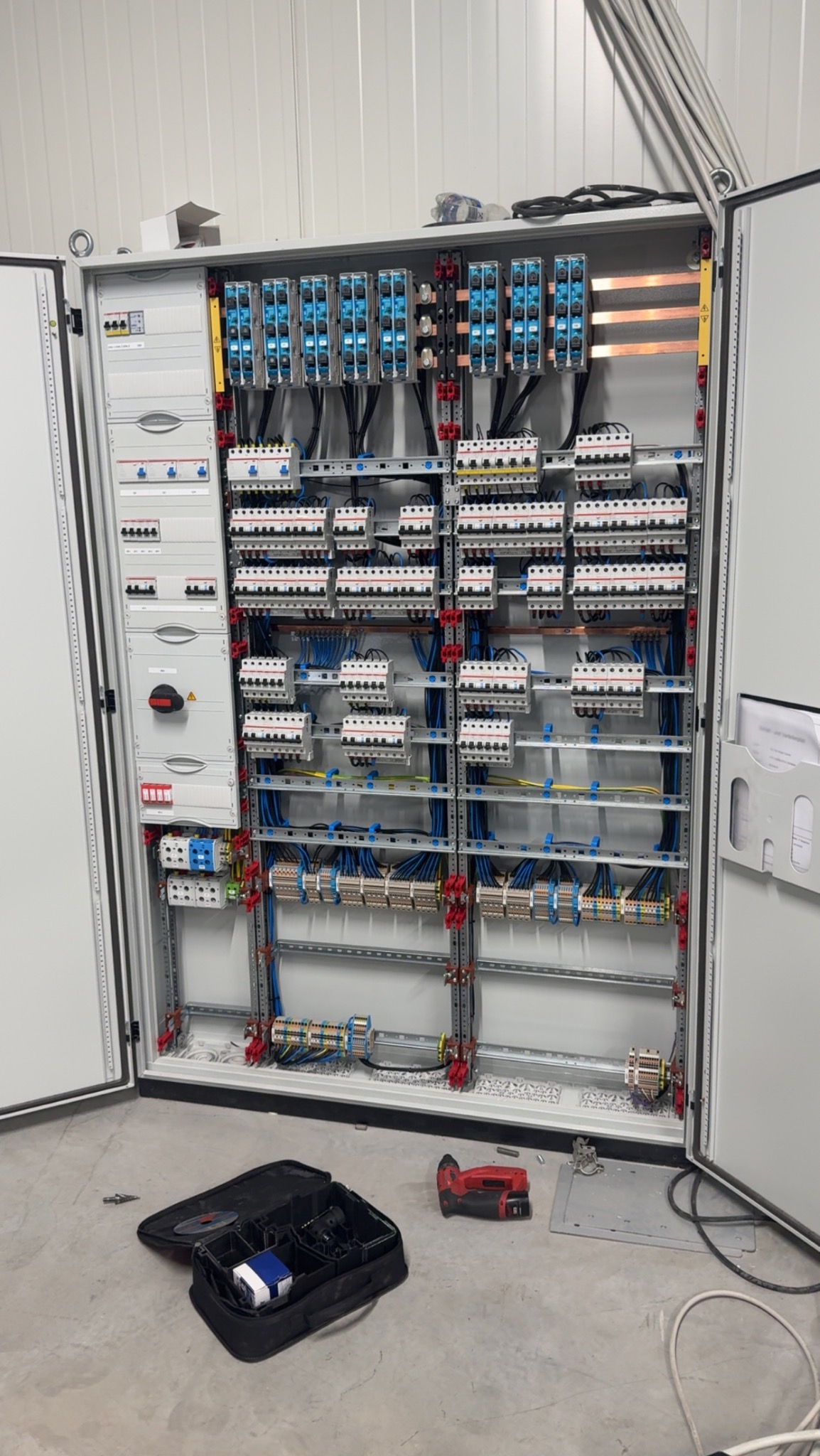 Otwarta szafa elektryczna z licznymi bezpiecznikami, przewodami i elementami montażowymi. Na podłodze narzędzia, w tym wiertarka i torba narzędziowa.
