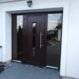 Brązowe drzwi wejściowe z szybami bocznymi, zamontowane w białej ramie. Widoczne narzędzia i taśma montażowa, sugerujące świeży montaż. Elegancki design.