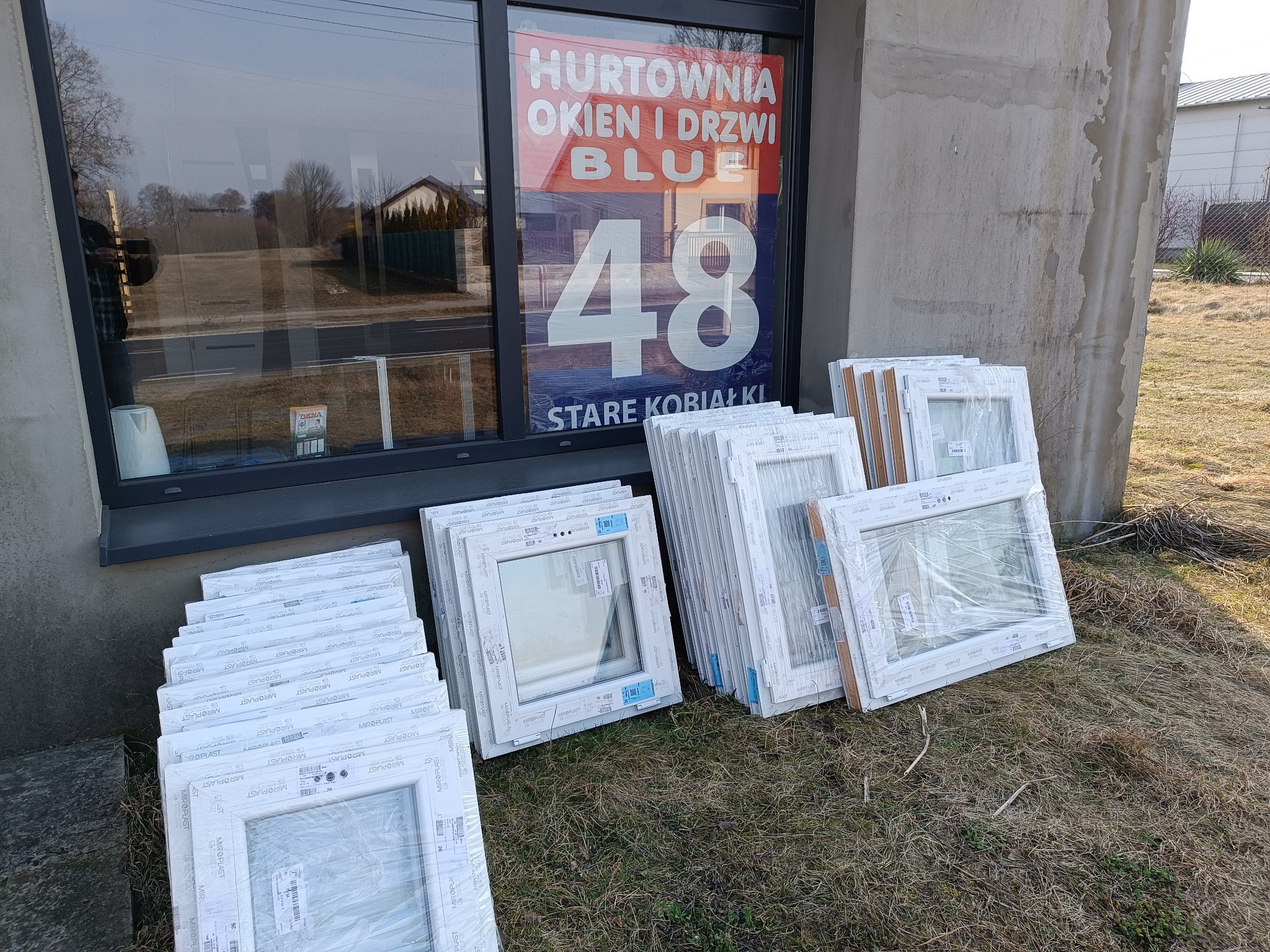 Stos okien PCV marki Miroplast, częściowo w folii, ustawionych przed oknem z reklamą hurtowni okien i drzwi. Widoczne budynki w tle.