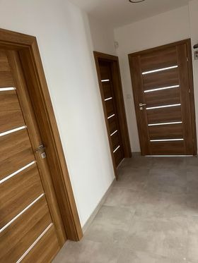 Wnętrze z trzema parami drzwi drewnianych o nowoczesnym designie, z poziomymi, jasnymi paskami. Drzwi w odcieniach brązu na tle białych ścian i szarej podłogi.