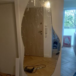 Jan Stanisławski - Montaż asymetrycznego lustra w przedpokoju, zabezpieczonego folią. Widoczne narzędzia i fragment podłogi w jodełkę. U góry kryształowy żyrandol.