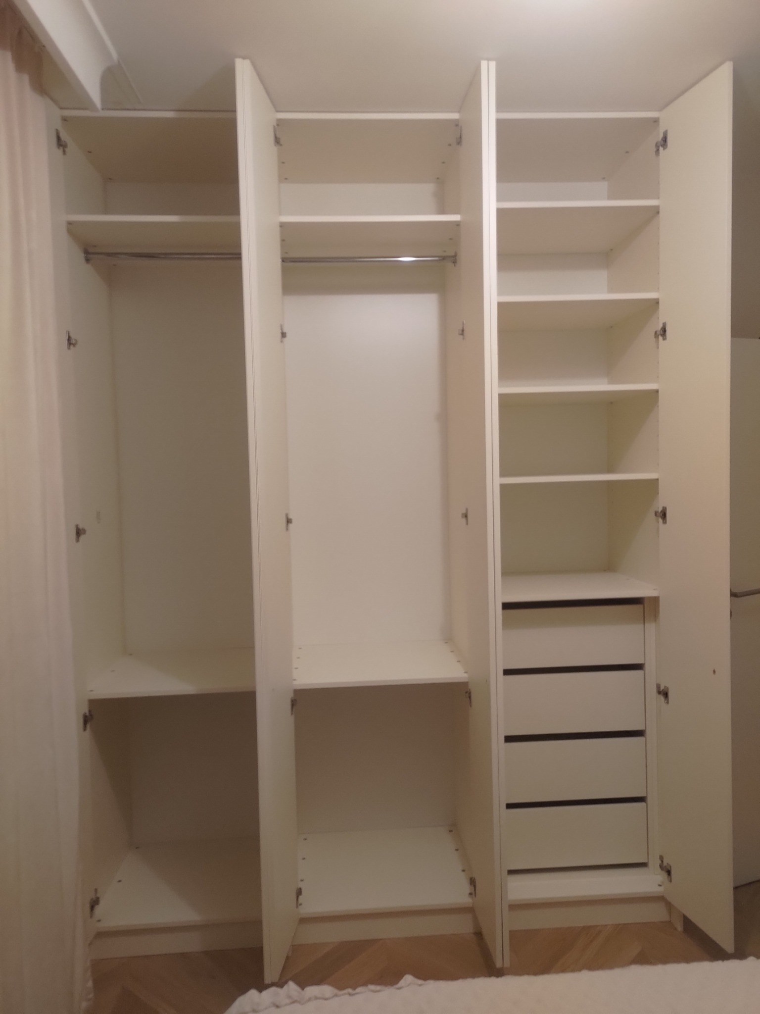 Biała, otwarta szafa wnękowa z półkami i szufladami, widok z przodu. Minimalistyczny design, jasne wnętrze, widoczne okucia. Szafa na podłodze z parkietu.