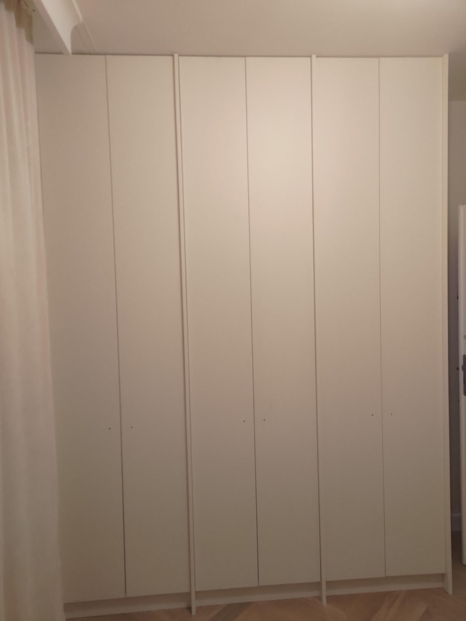 Biała, zabudowana szafa z sześcioma drzwiami, widoczna z bliska, z delikatnym oświetleniem i fragmentem zasłony po lewej stronie. Minimalistyczny design.