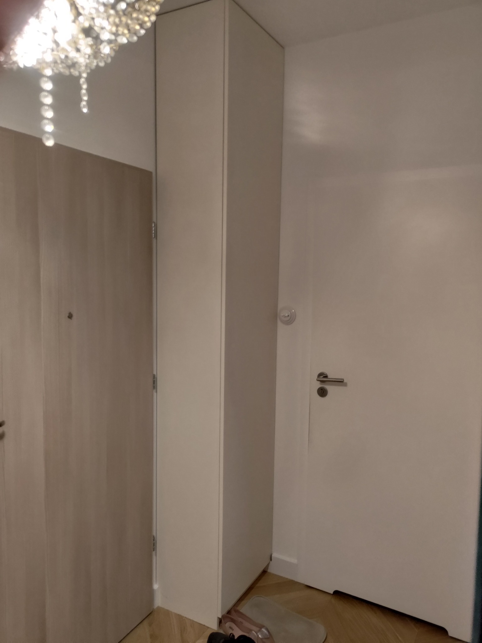 Wnętrze: zabudowa szafy wnękowej z jasnego drewna, widoczna część sufitu z ozdobnym żyrandolem, drzwi wejściowe. Minimalistyczny design, jasne kolory.