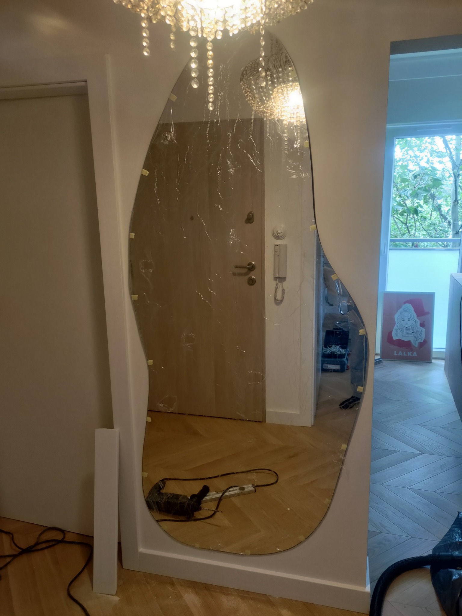 Montaż asymetrycznego lustra w przedpokoju, zabezpieczonego folią. Widoczne narzędzia i fragment podłogi w jodełkę. U góry kryształowy żyrandol.