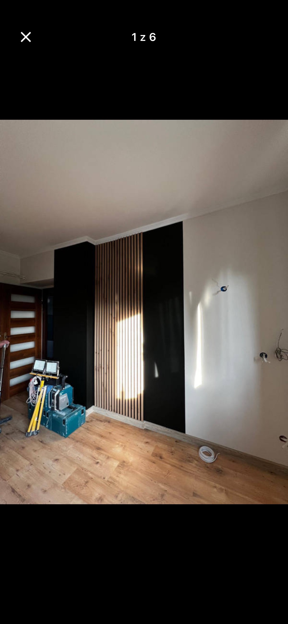 Nowoczesne wykończenie ściany z drewnianymi lamelami i czarną, błyszczącą płytą. Na podłodze panele. Widoczne narzędzia i niedokończone instalacje elektryczne.