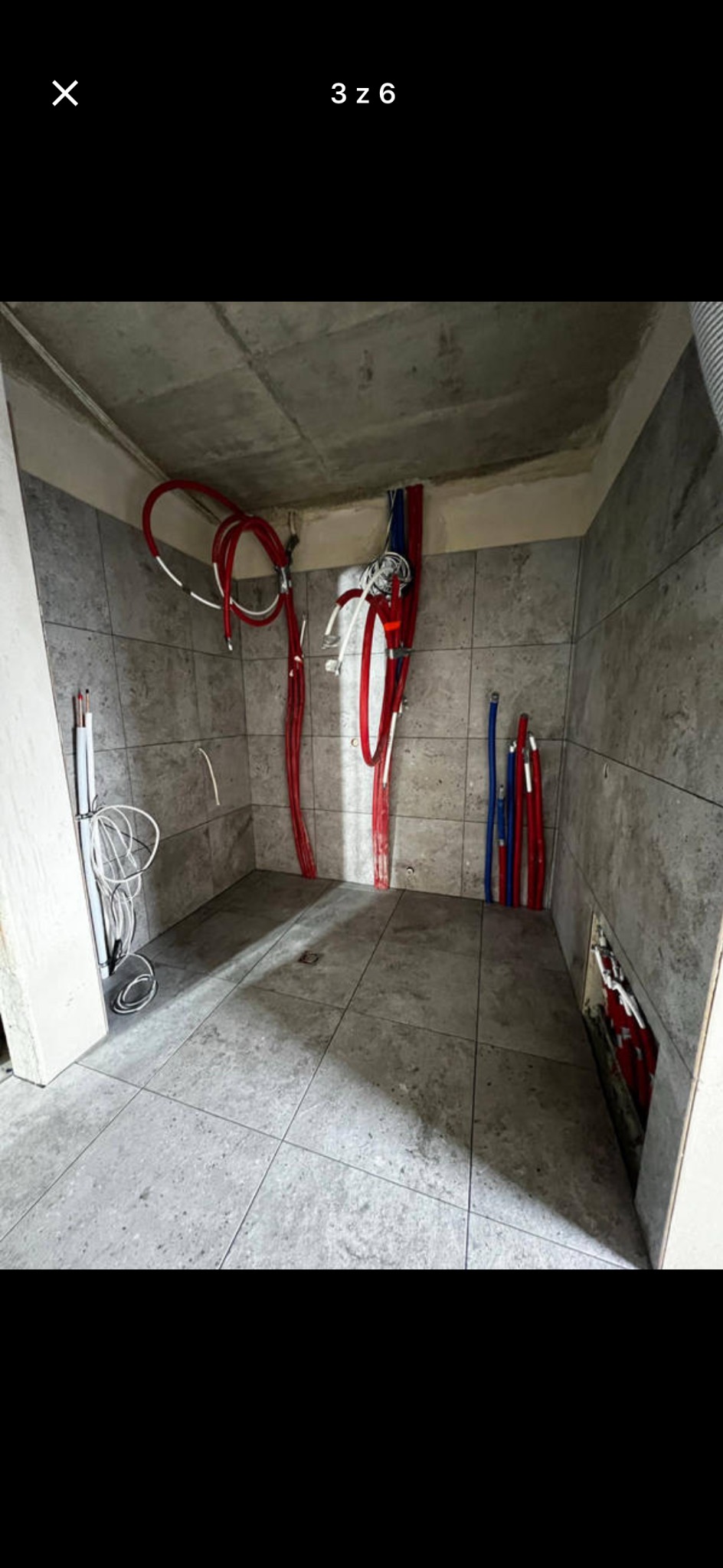Wnętrze łazienki w trakcie remontu: szare płytki na ścianach i podłodze, widoczne rury instalacji wodnej (czerwone i niebieskie). Surowy betonowy sufit.