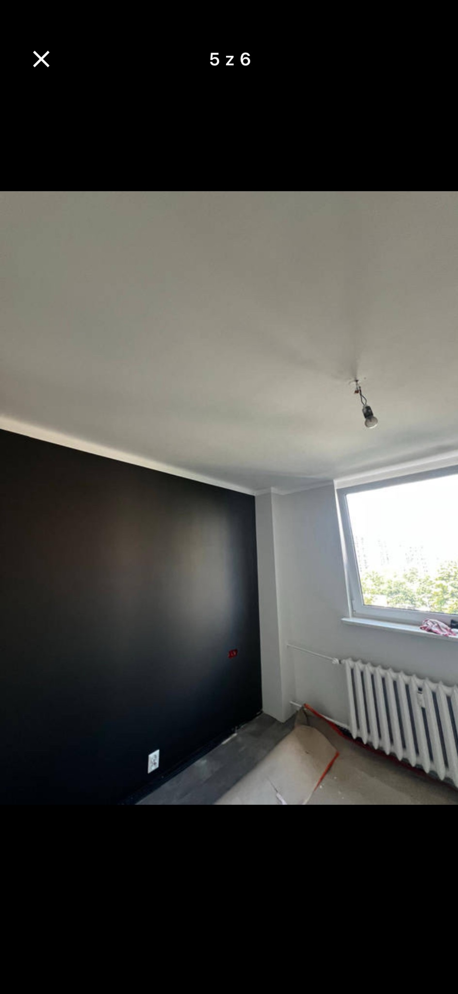 Pokój po malowaniu: czarna ściana kontrastuje z białą resztą. Widoczne gniazdka, okno z grzejnikiem i zabezpieczona podłoga. Minimalizm i nowoczesność.