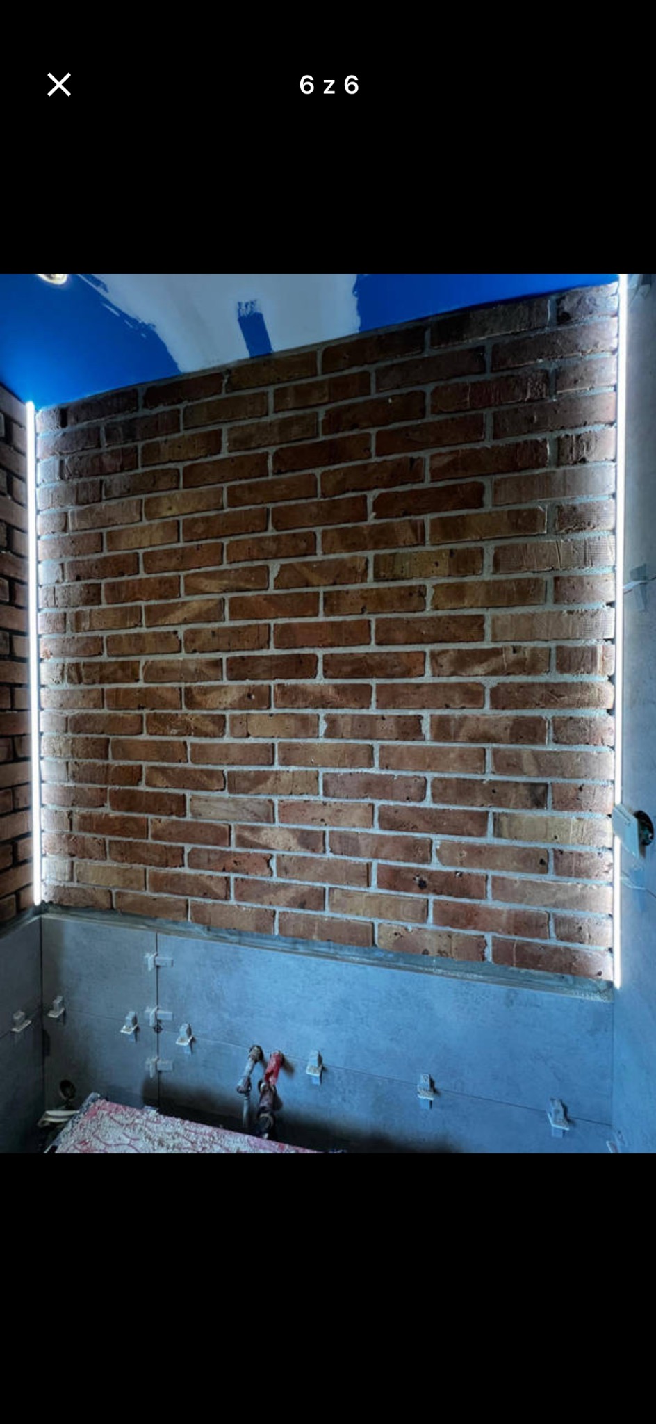 Ściana z cegły klinkierowej z oświetleniem LED na krawędziach, płytki w trakcie montażu na dolnej części ściany, niebieskie malowanie sufitu w tle.