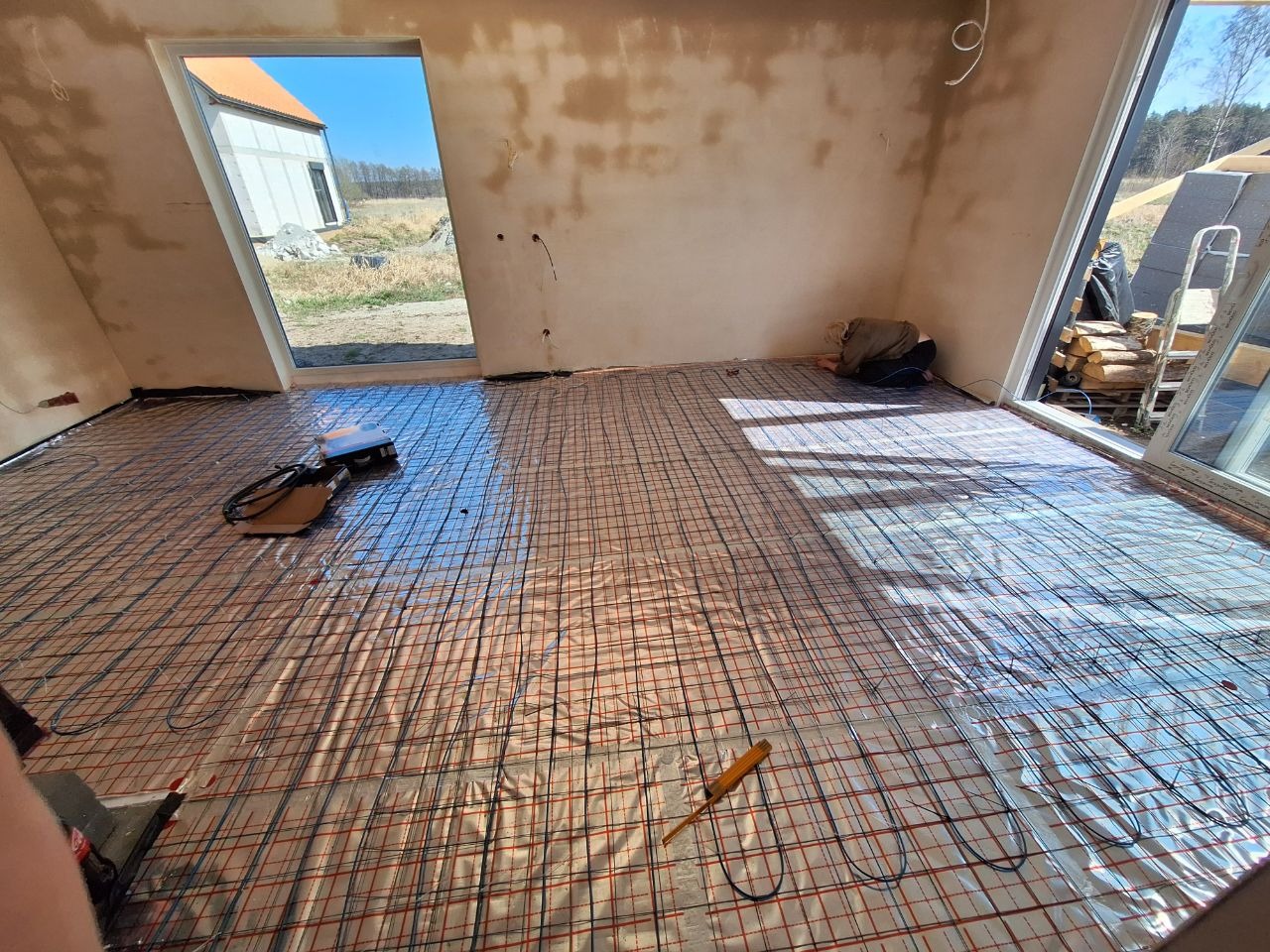 Rozłożona mata grzewcza na podłodze w surowym wnętrzu. Widoczne przewody, narzędzie i osoba pracująca przy instalacji. W tle okno z widokiem na budynek i drzewa.