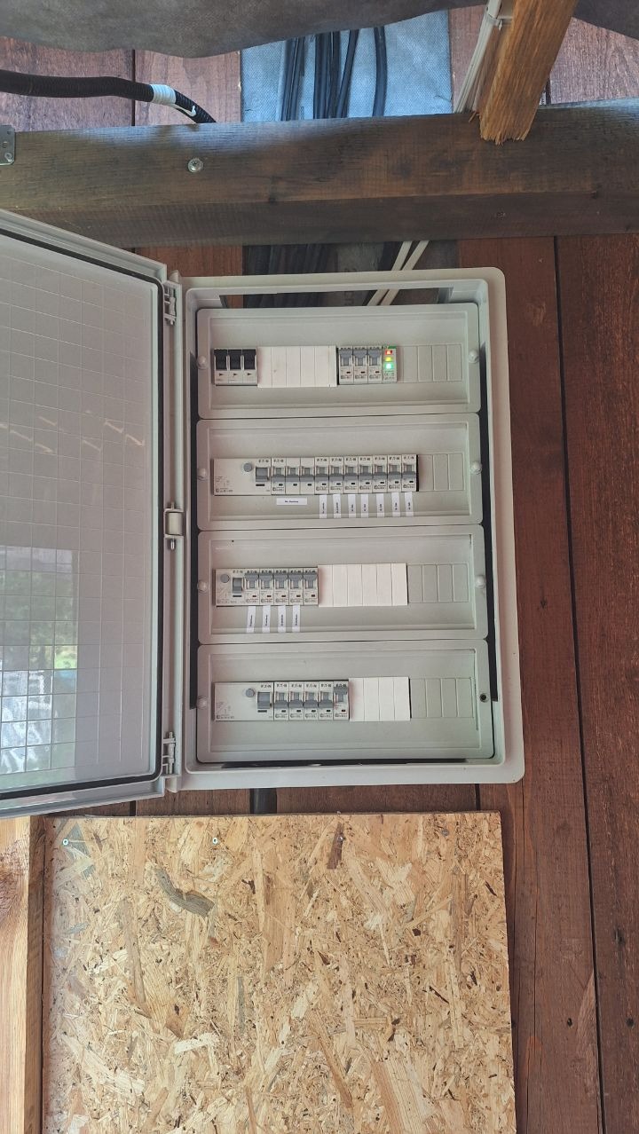 Otwarta skrzynka rozdzielcza z bezpiecznikami na drewnianej ścianie. Widoczne kable i elementy instalacji. Oświetlenie sufitowe.