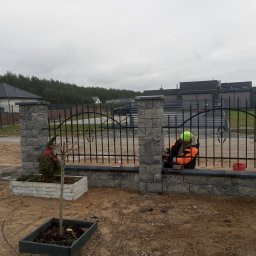 Projekt Ogrod - Montaż ozdobnego ogrodzenia z kamiennych bloczków i metalowych przęseł. Pracownik w odblaskowej kamizelce przy pracy. W tle domy i drzewa.