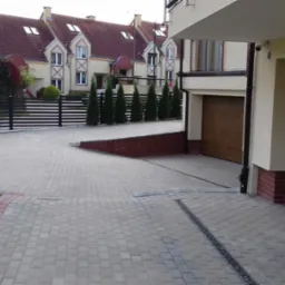 Ułożona kostka brukowa na podjeździe do garażu, widok na nowoczesny dom z brązowymi bramami garażowymi i rząd tuj przy ogrodzeniu.