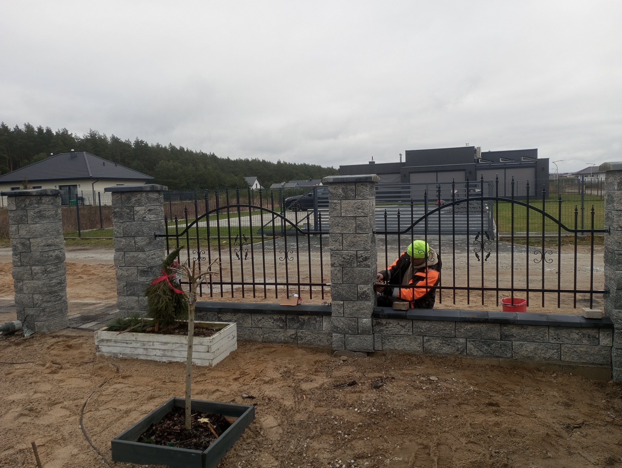 Montaż ozdobnego ogrodzenia z kamiennych bloczków i metalowych przęseł. Pracownik w odblaskowej kamizelce przy pracy. W tle domy i drzewa.